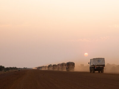 WFP/Samantha Reinders A convoy of trucks drives on a dusty road at sunset, the sun low on the horizon casting long shadows under a clear sky.