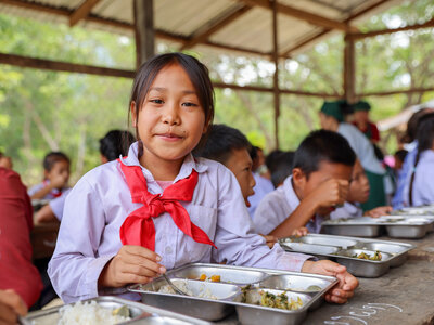 Photo: WFP/Vilakhone Sipaseuth Students sit together at long wooden tables in an open-air school dining area, eating a meal served in metal trays. A student in the foreground holds a spoon while other students eat and talk in the background, illustrating a school meals programme.