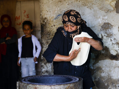Photo: WFP/Ahmed Basha A person in a patterned headscarf stretches dough over a cooking surface, with two others standing in the background against a textured wall.