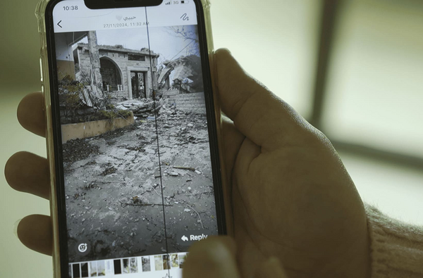 Hand holds mobile phone screen showing a destroyed house with a bulldozer in the background