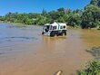 A small amphibious vehicle with large, partially submerged tires sits near a bushy bank by brown water, its front hatch open.