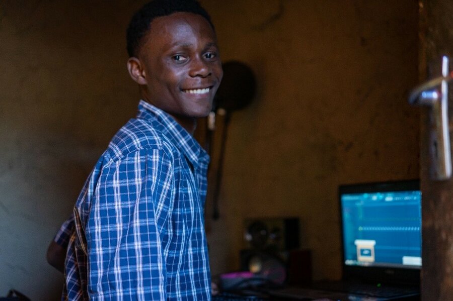 A boy smiles at the camera with a computer monitor in the background.