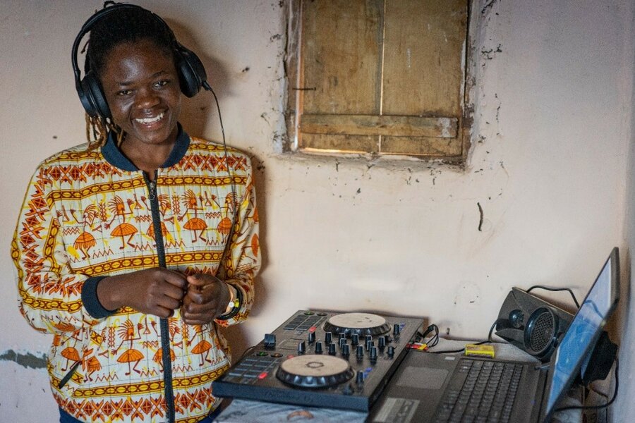 A woman wearing headphones stands next to a soundboard and laptop.