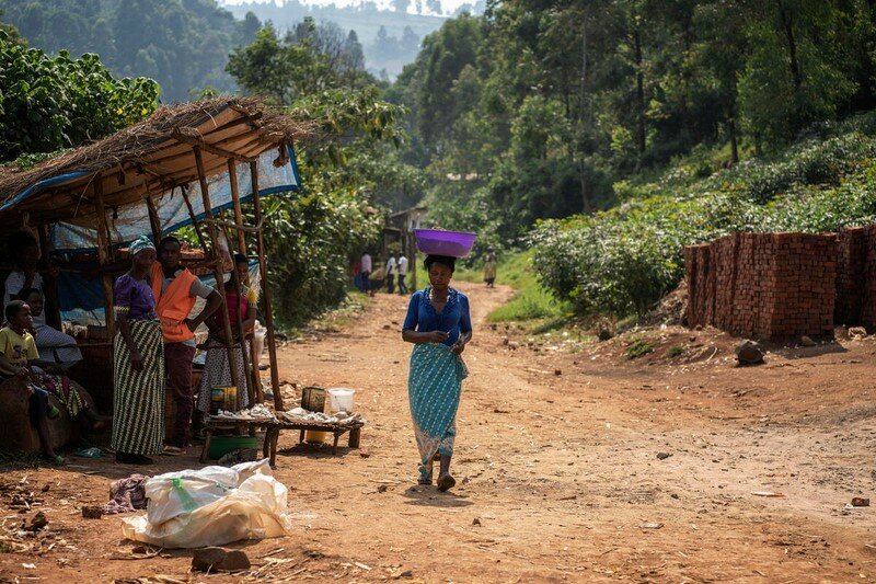 A woman walks on a dirt road with a dish on her head.