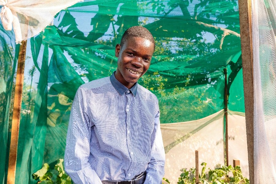 A boy in a blue button down shirt stands smiling in front of green netting.