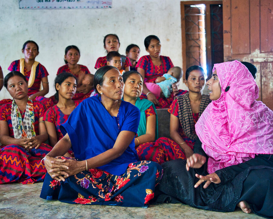 a group of tribal women in Bangladesh sit on the floor talking to a woman in a pink headscarf