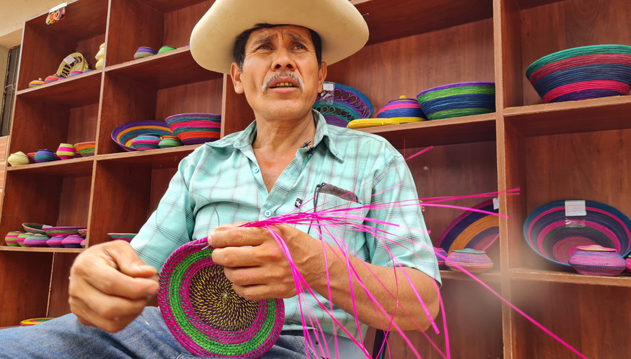Bolivian Indigenous man in a straw hat weaves a basket using coloured wicker strands