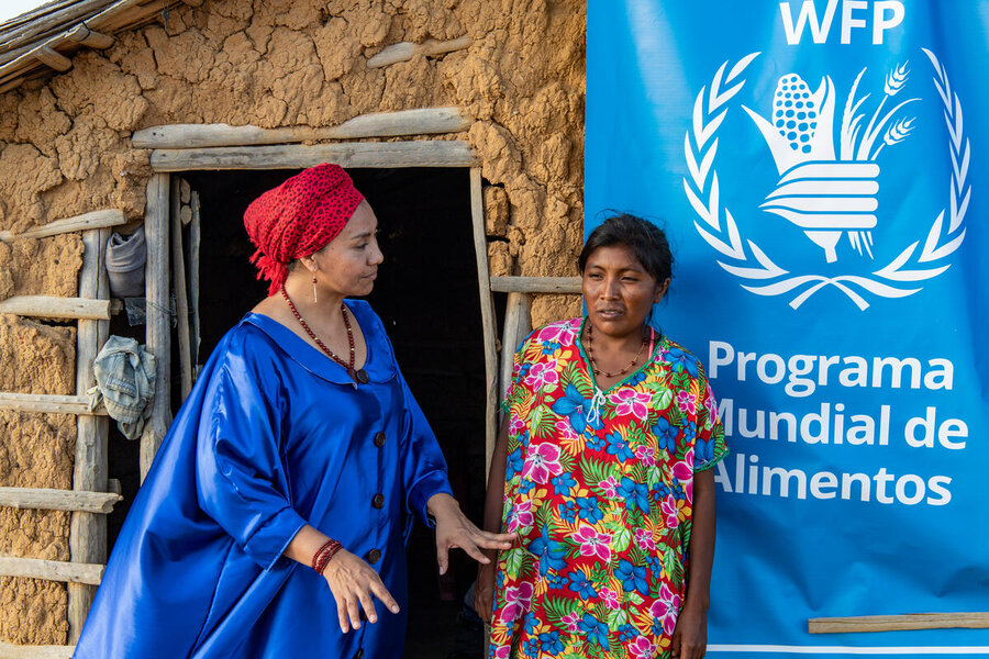 WFP Indigenous staff member in a blue cape and red headdress speaks to woman in flowery dress outside a mud hut with WFP banner