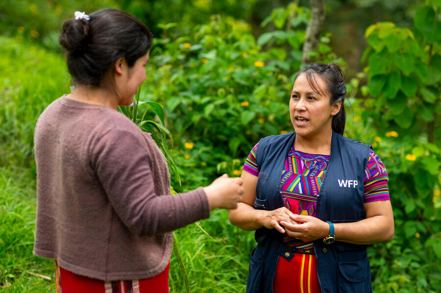 Indigenous woman in traditional dress and navy blue WFP vest speaks to a woman who turns her back to the camera against forest background in Guatemala