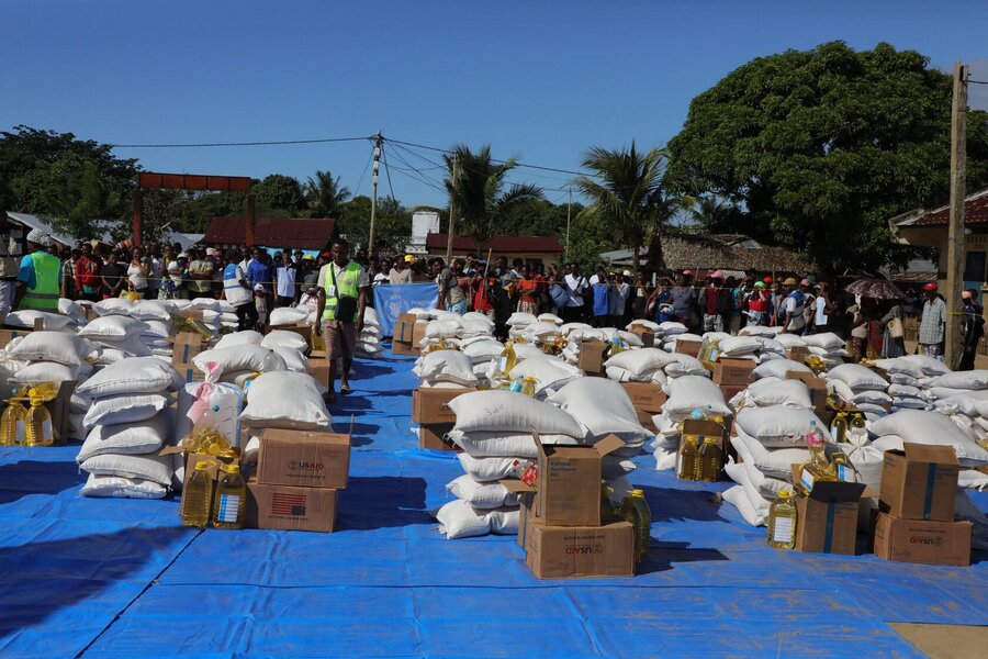 Rows of food sacks and food assistance boxes on blue sheeting as people look on in the background