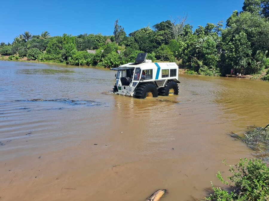 A small amphibious vehicle with large, partially submerged tires sits near a bushy bank by brown water, its front hatch open.