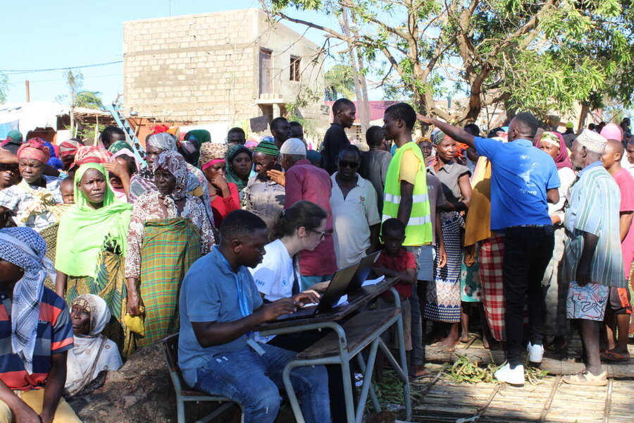 Two WFP workers at laptops in the outdoors amid a crowd of people lining up