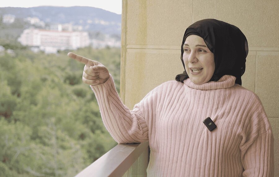 A young woman in headscarf on a balcony points outwards - there is greenery in the background