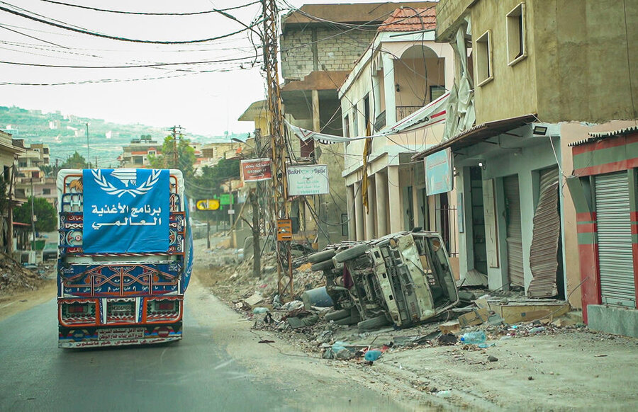 A colourful truck - draped with a WFP logo banner in Arabic - makes its way up a deserted road past an burnout, overturned car 