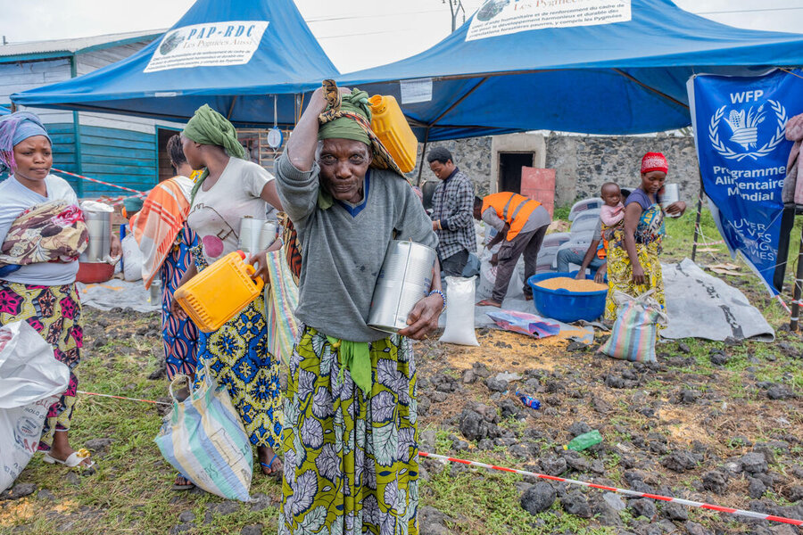 A woman at a WFP distribution sight looks at the camera holding a yellow jerrycan over her head and a big USAID tin