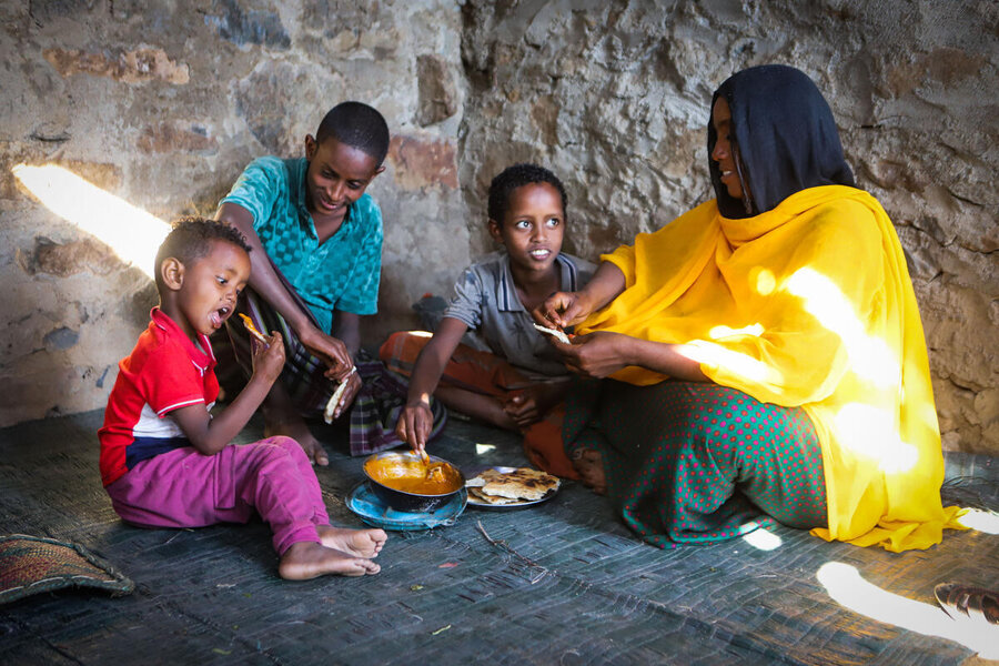  A woman in a yellow shawl observes as three boys sit on the floor against stone walls, eating a meal.