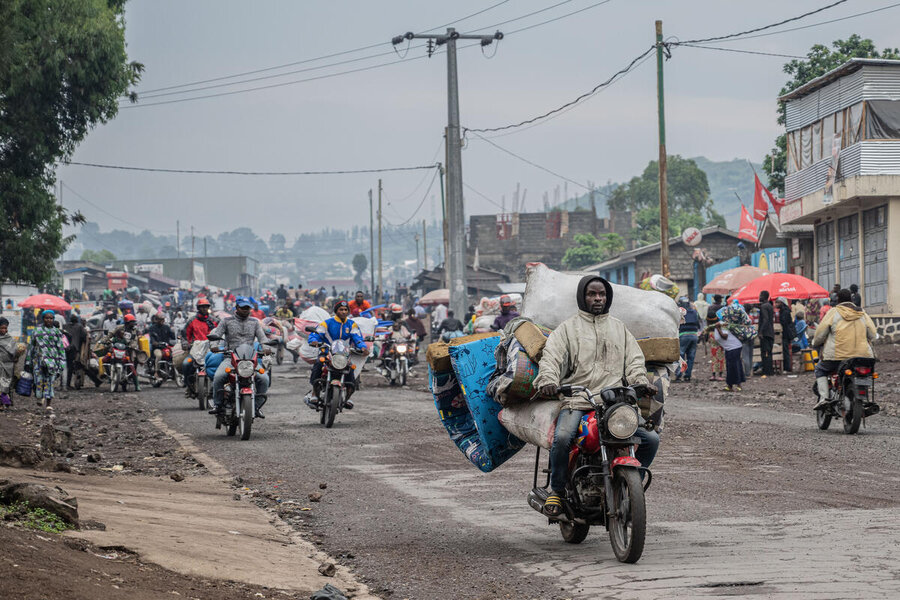 A man rides a heavily loaded motorbike through a crowded street, carrying mattresses and belongings, as others follow