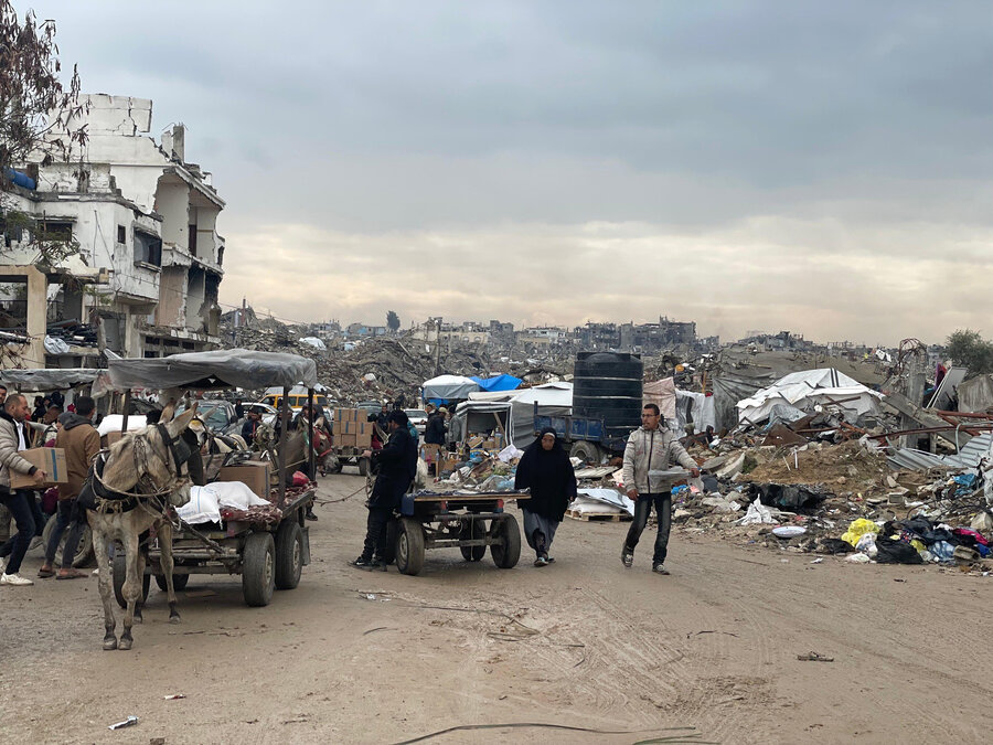 A wrecked street in Gaza, with collapsed buildings and debris stretching behind. Residents move through the ruins, some on donkey carts, others on foot, amid makeshift shelters and grey skies