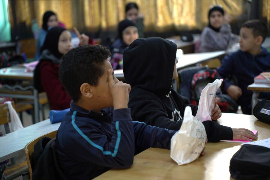 A boy next to another boy in class looks away from the camera clutching a white packet