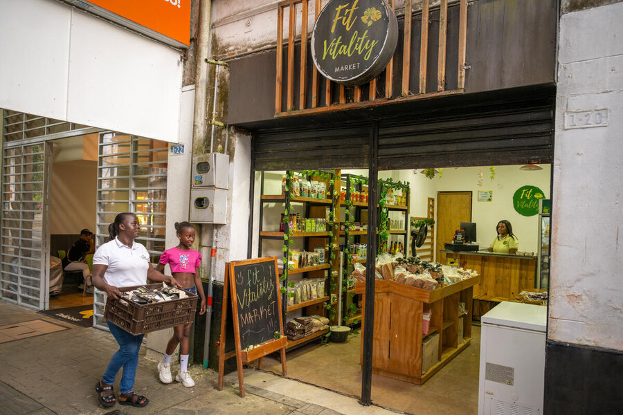 A woman carrying a box full of potato chips packs and her daughter approach a shop called Fit Vitality