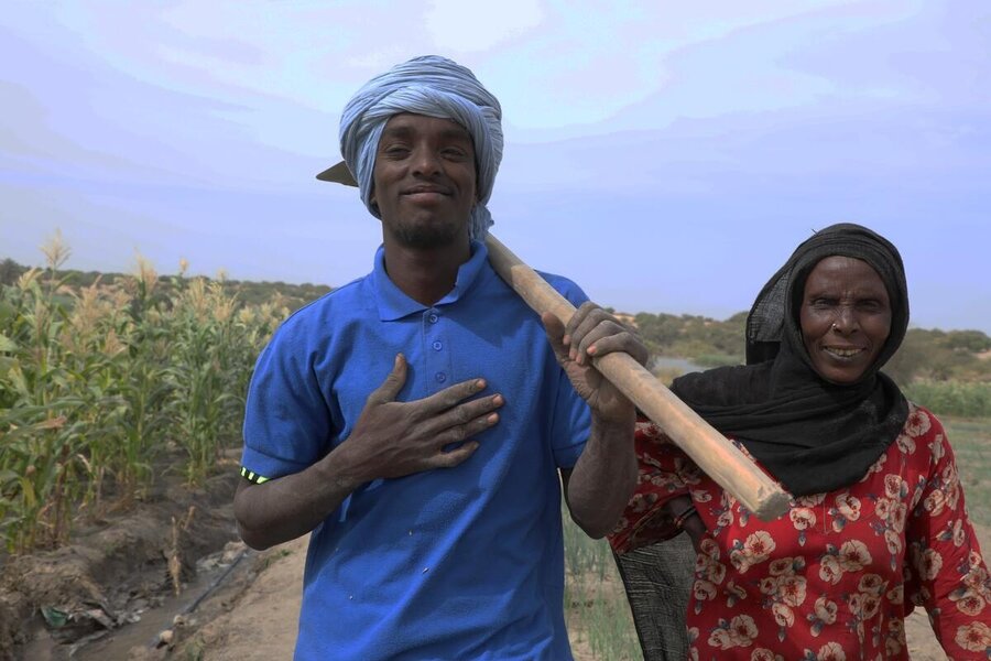 Two smiling individuals stand in a field, one holding a farming tool over their shoulder.
