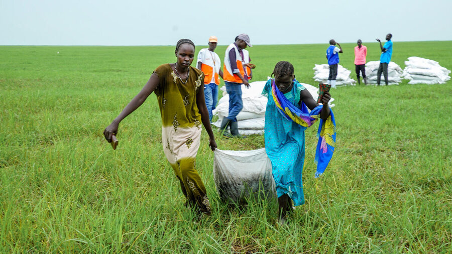 Two people carry a heavy sack across a grassy field while others work in the background.
