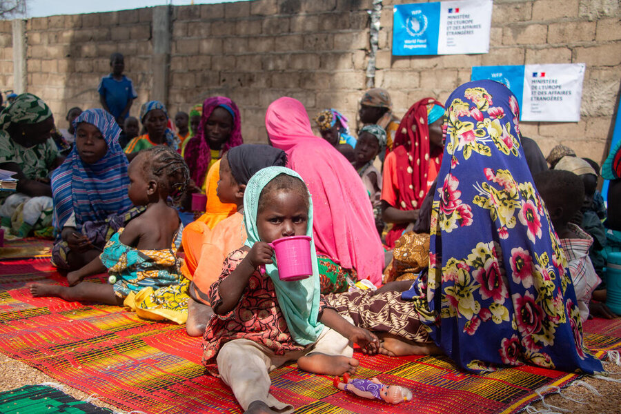 Children drink from cups on colourful mats while others gather outside near brick walls.