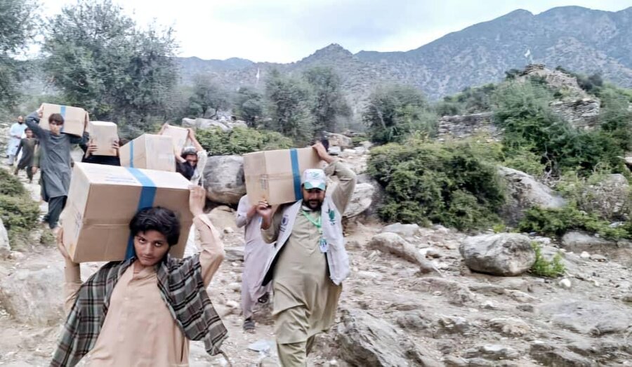 People carry large boxes on their heads through rocky terrain with mountains behind.