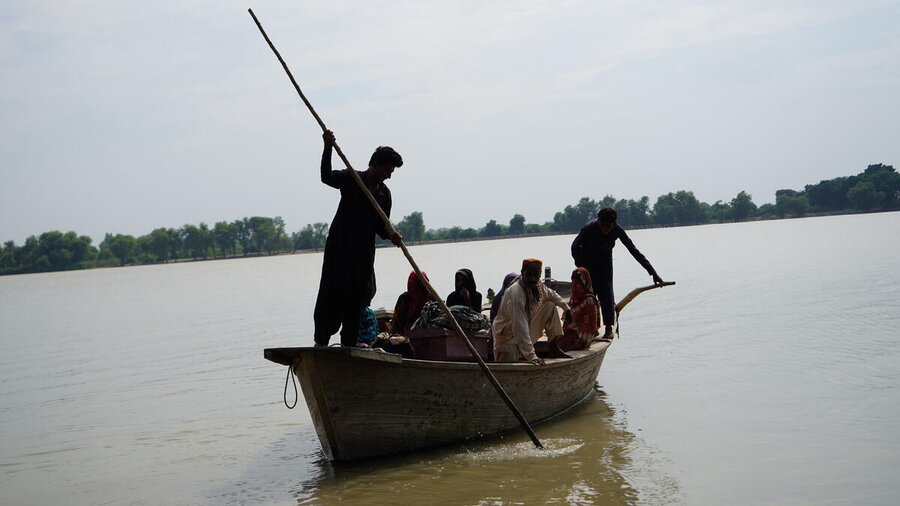People travel in an open boat steered by poles under cloudy skies near tree-lined shores.