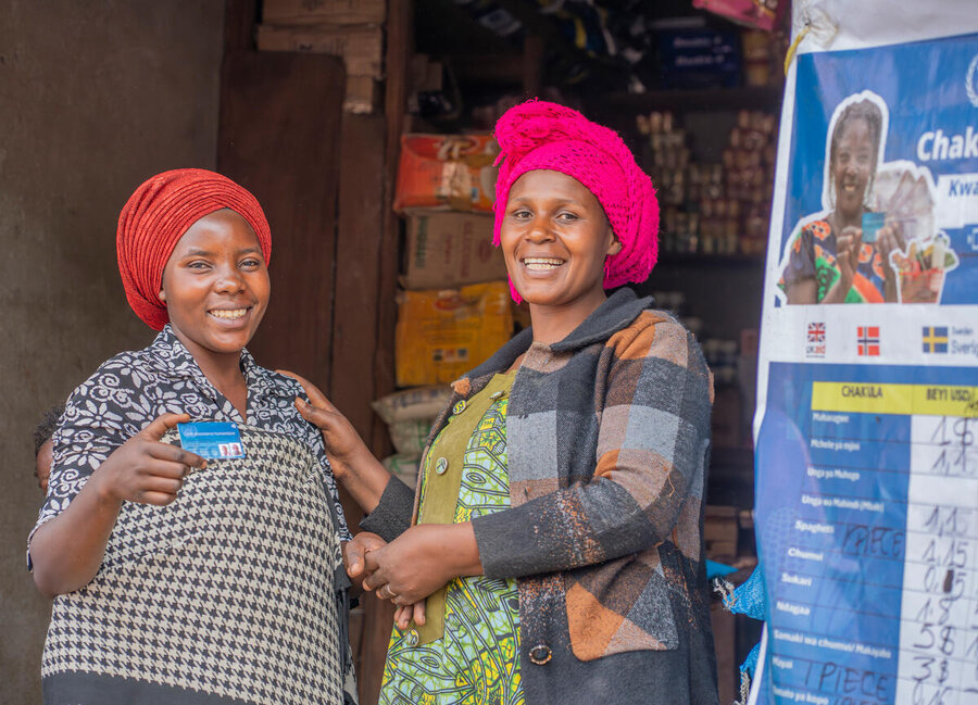 Two people smiling in front of a shop; one holds an ID card. Shelves and a poster with flags and prices are visible behind them.