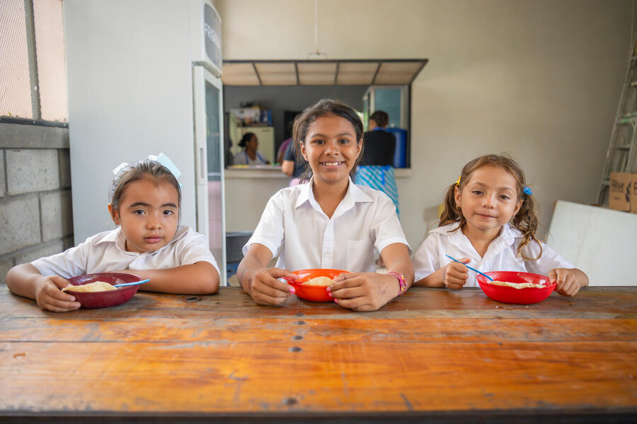 Three children in white shirts eat from bowls at a wooden table, smiling at the camera.